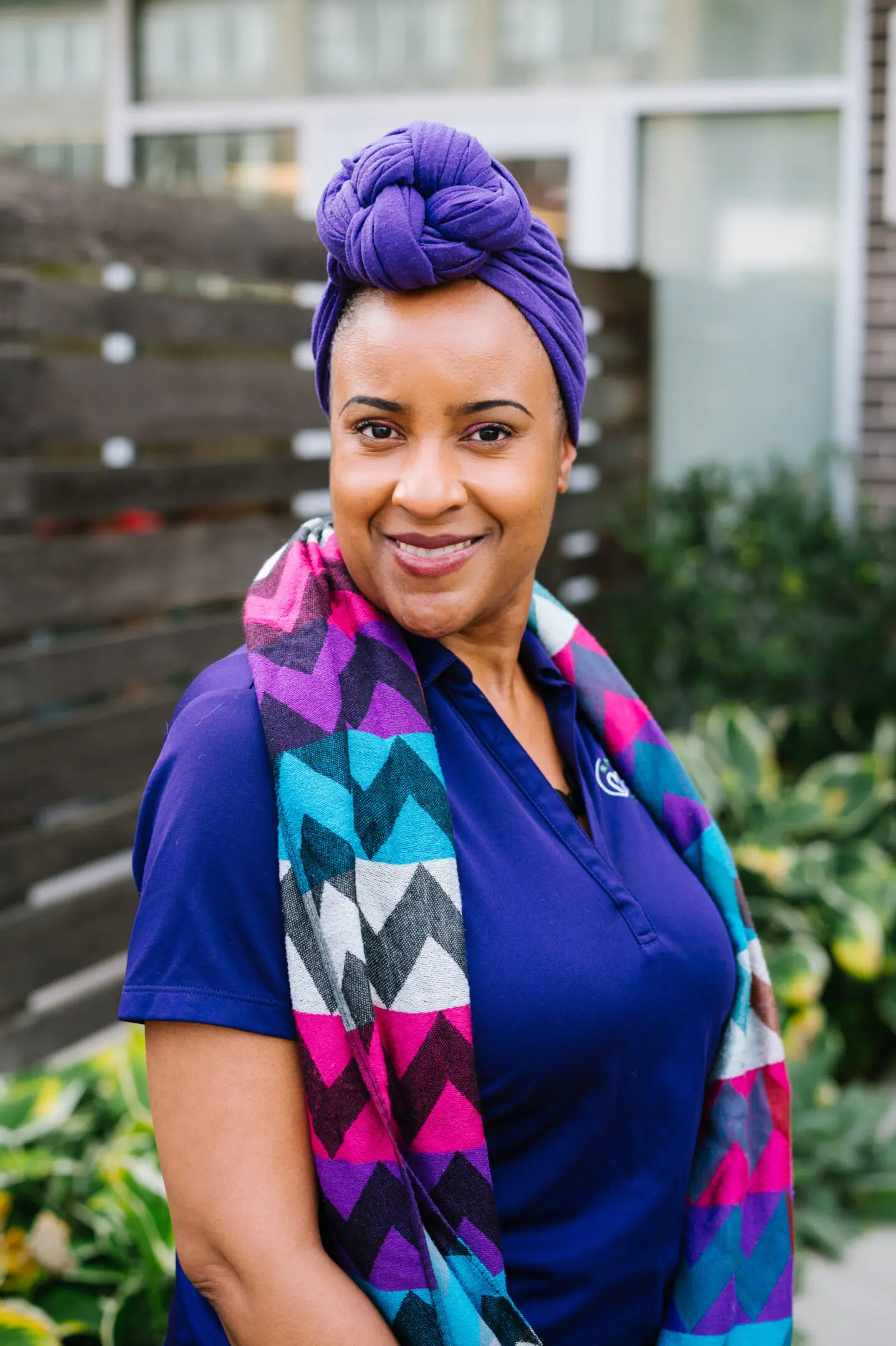 Smiling woman wearing a purple headwrap and colorful scarf.