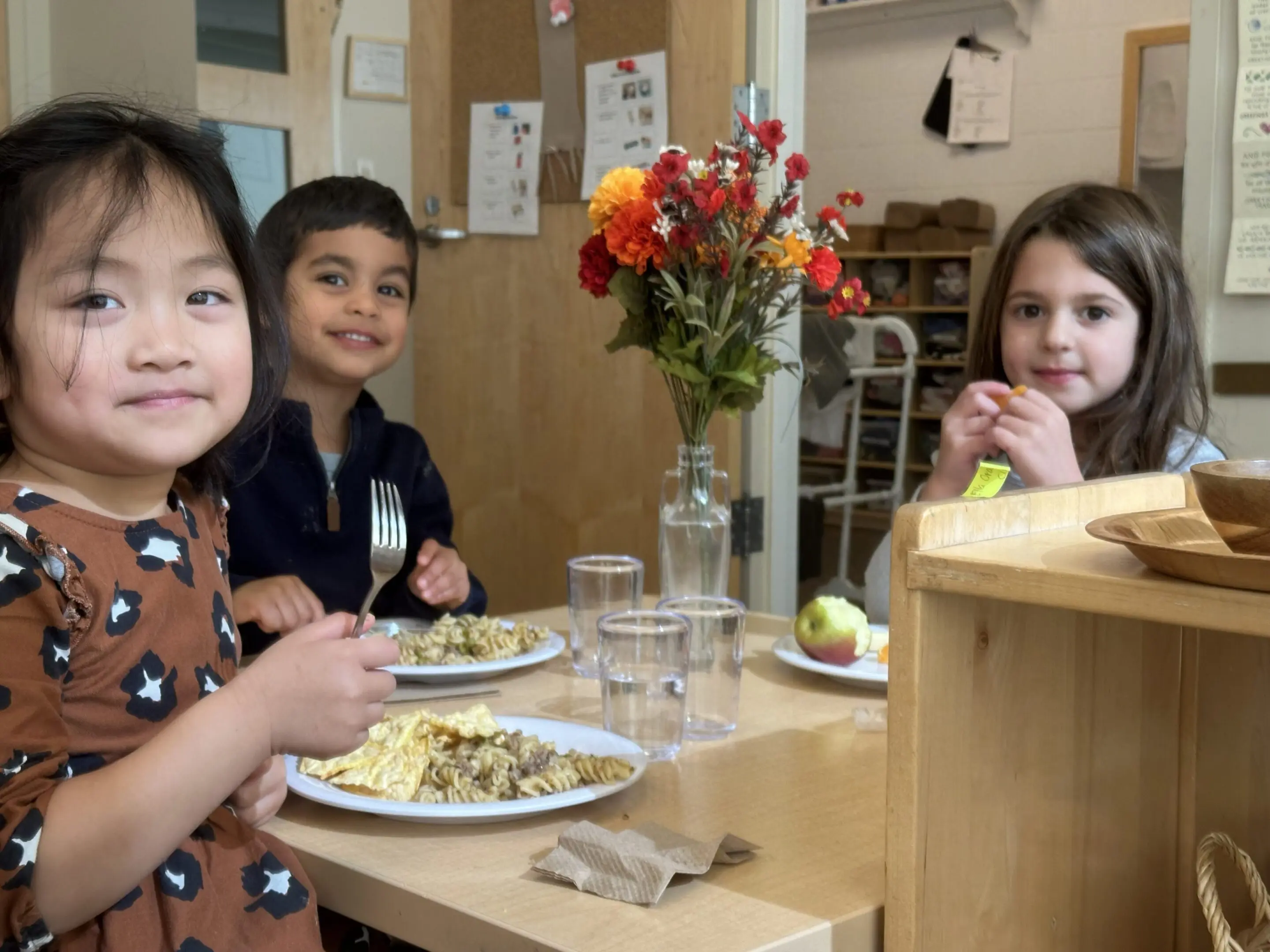 Three children sitting at a table with food and flowers in the center.