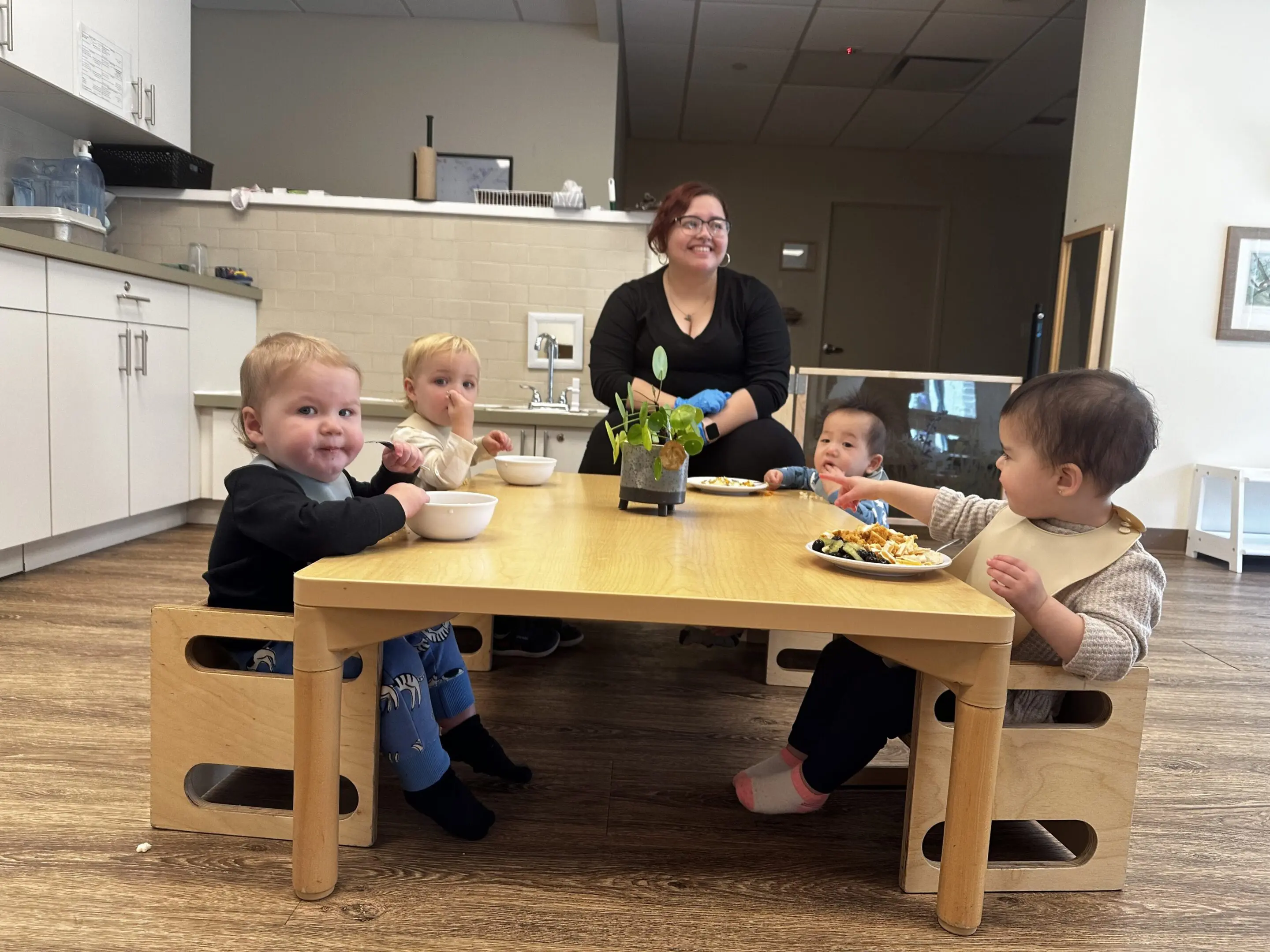 Four toddlers sitting around a low table with a caretaker smiling behind them.