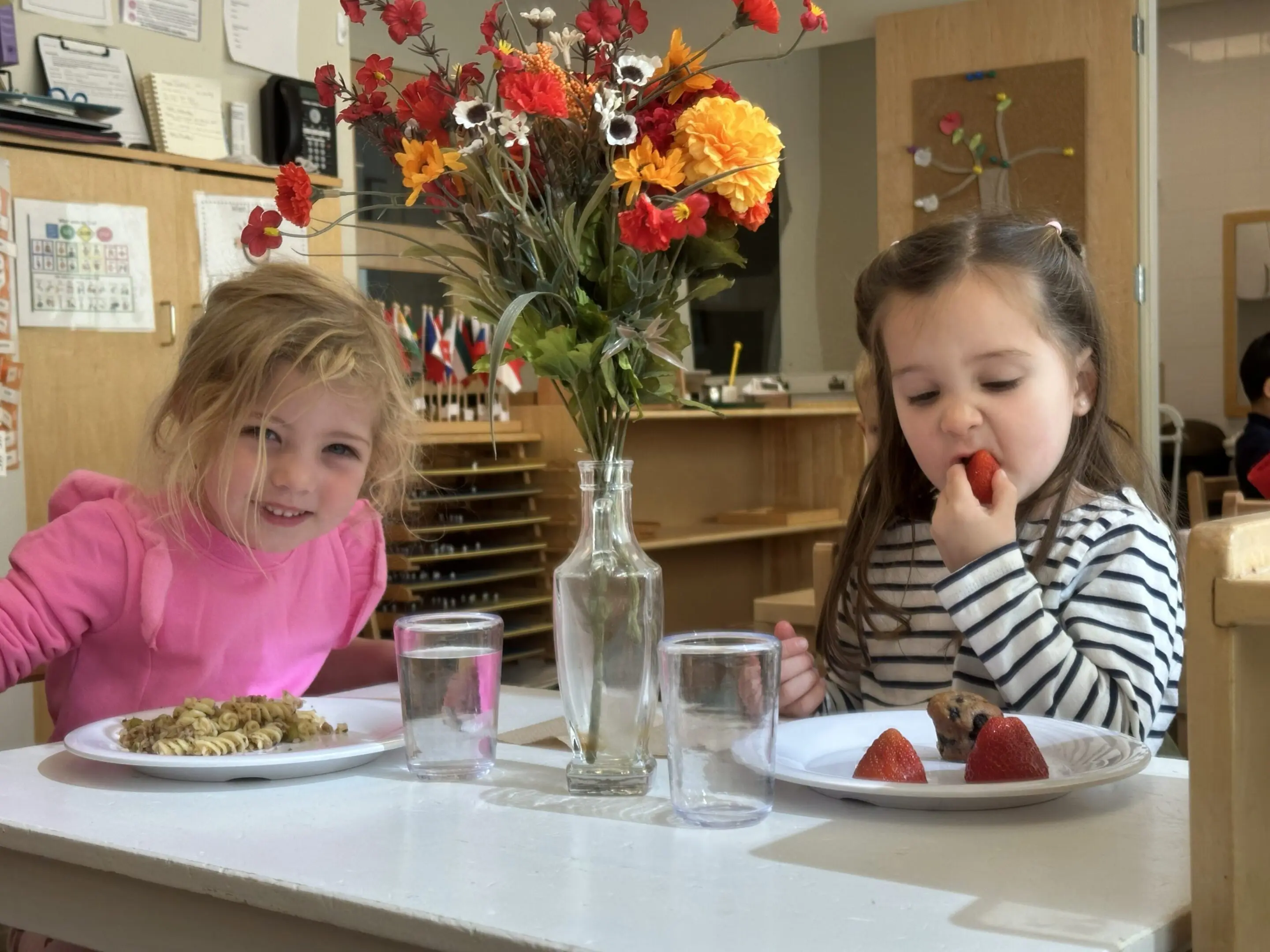 Two young girls enjoying snacks at a kitchen table with flowers.